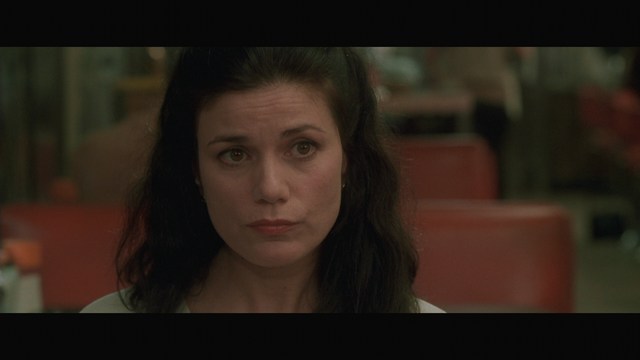 A close-up of a woman with dark hair and a serious expression sitting in a diner, with blurred background elements suggesting a busy atmosphere.