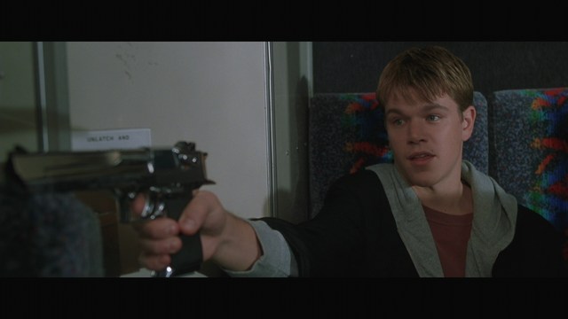 A young man aiming a handgun while seated, wearing a hoodie and looking tense.