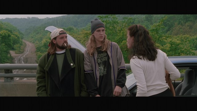 Scene featuring three characters on a roadside, discussing animatedly. A green landscape is visible in the background.