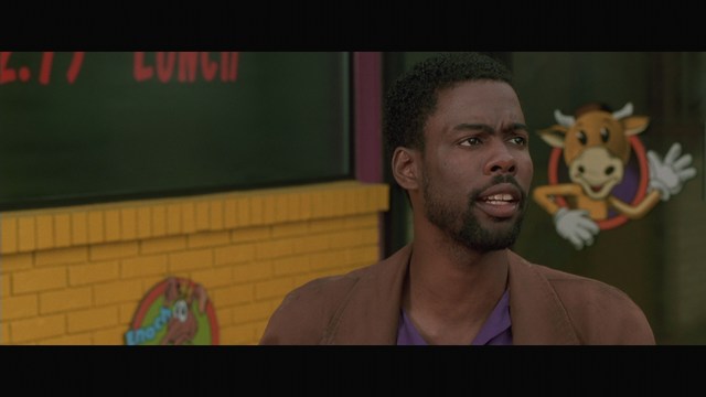 A man with short hair wearing a brown jacket and purple shirt, speaking animatedly in front of a colorful restaurant featuring a cartoon cow logo in the background.