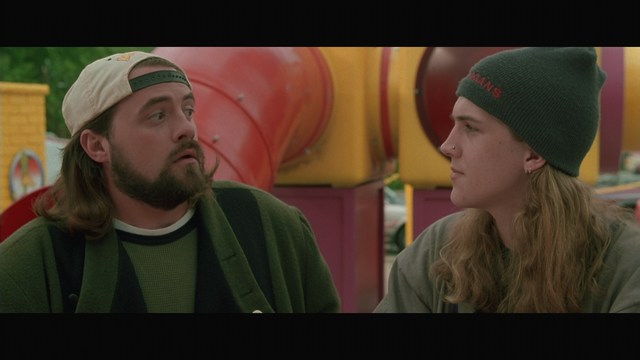 Two men engaged in conversation in a scene from a movie, set against a colorful playground background.