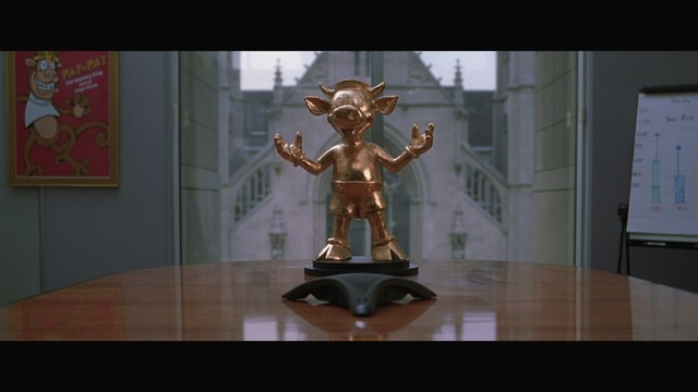 A bronze trophy figure of a cartoonish character with arms outstretched, positioned on a wooden table in a corporate setting, with a large window showcasing a mansion in the background.