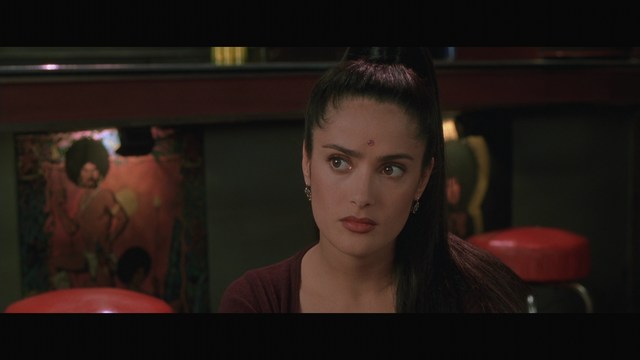 A close-up shot of a woman with long dark hair, wearing a maroon top and earrings, sitting in a dimly lit setting with red stools and decorative artwork in the background.