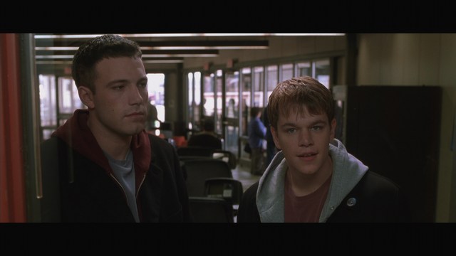 Two male characters in a movie scene, standing indoors near a doorway, with a blurred background of a waiting area.
