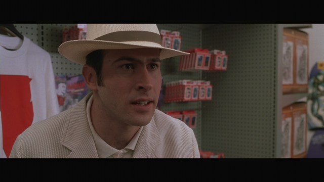 A man wearing a white suit and a fedora hat, looking surprised or upset, stands in a store with various products displayed in the background.
