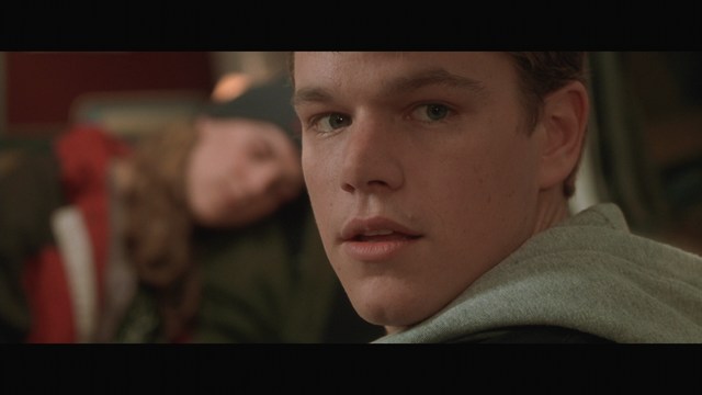 Close-up shot of a young man with short hair, wearing a gray hoodie, looking over his shoulder with a contemplative expression, while another person is asleep in the background.
