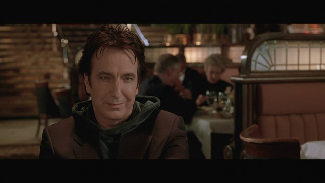 A man with dark hair and a hooded jacket smiles while sitting at a restaurant table, with blurred diners in the background.