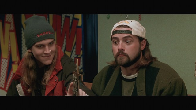 Two male characters in a cozy indoor setting, one with long hair and a beanie, smirking while holding a small object, and the other with a beard and cap, looking surprised. A colorful background with posters can be seen.