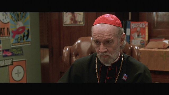A close-up of a man wearing a red cardinal's hat and black clerical robes, seated in a leather armchair with a serious expression, in an office setting adorned with colorful posters.