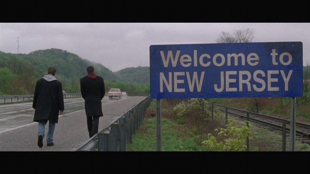 Two figures walking along a highway next to a 'Welcome to New Jersey' sign, with greenery in the background.