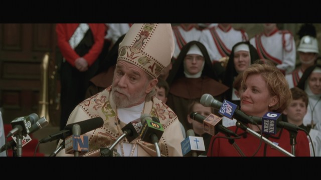 A scene from a movie featuring a man dressed in a bishop's robe speaking at a podium surrounded by microphones, with a woman in a red outfit smiling next to him, and an audience in the background.