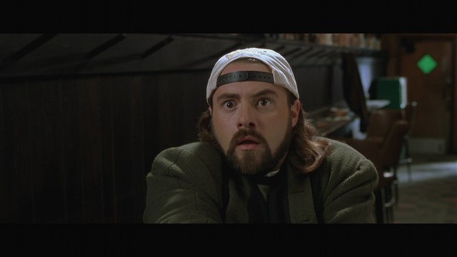 A man with long hair and a baseball cap appears shocked or surprised, sitting in a dimly lit room with wooden paneling and chairs in the background.