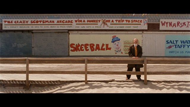 A still from a movie featuring a man standing on a boardwalk in front of an arcade with signs for Skeeball, saltwater taffy, and various attractions.