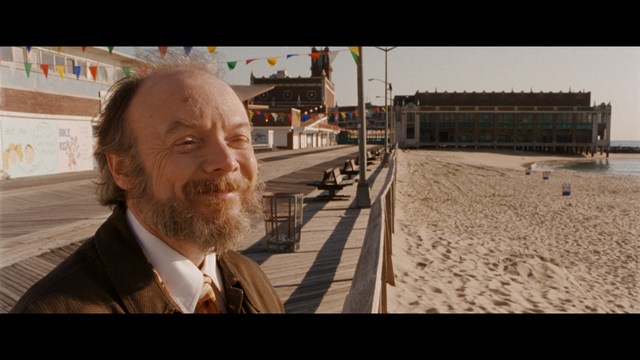 A man with a beard and mustache smiles warmly while standing on a boardwalk by the beach, with colorful flags overhead and a large building in the background.