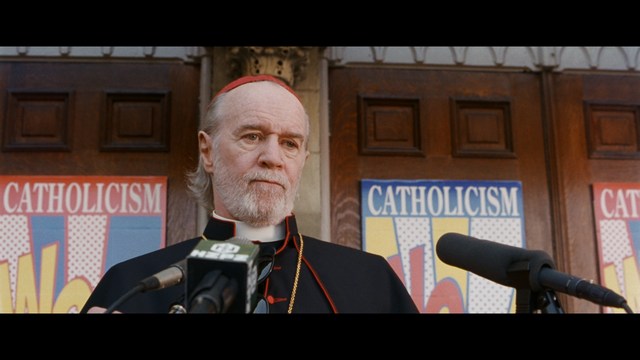 A serious-looking cardinal stands in front of a wooden backdrop with colorful posters related to Catholicism, holding a microphone at a press conference.