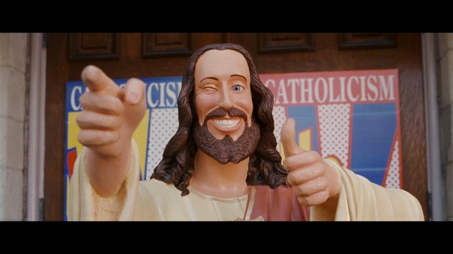 Statue of a smiling figure resembling Jesus, giving a thumbs up gesture, set against a colorful backdrop with signs related to Catholicism.