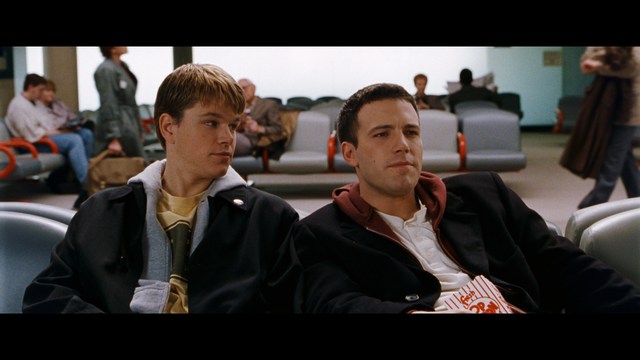 A scene from a movie featuring two young men sitting closely together in an airport waiting area, with one holding a box of popcorn.