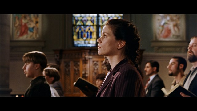 A group of people standing in a church, with a focus on a woman in a striped blazer looking up, while others are holding books or looking ahead.