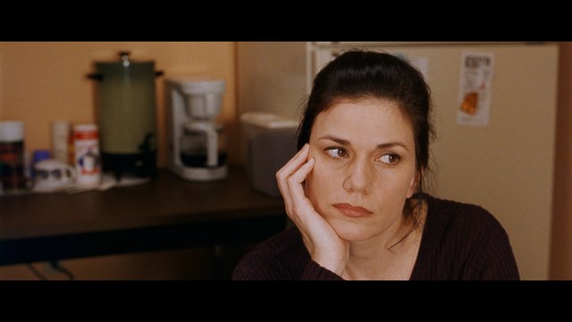 A woman with a pensive expression rests her chin on her hand, sitting at a table with kitchen appliances and items in the background.