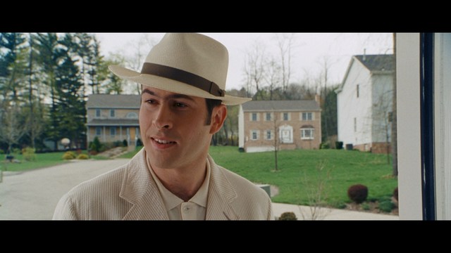 A man in a white suit and hat stands at a doorway, smiling, with a suburban landscape in the background.