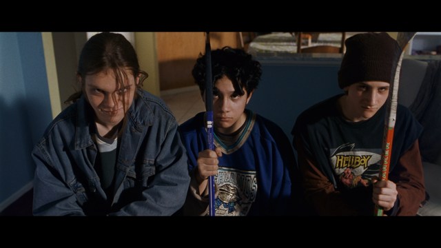 Three young individuals sitting together, two holding hockey sticks, with a casual and intense expression in a domestic setting.