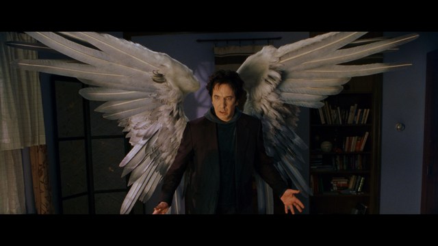 A man with large, feathered angel wings stands in a room, wearing a black suit and looking directly at the viewer.
