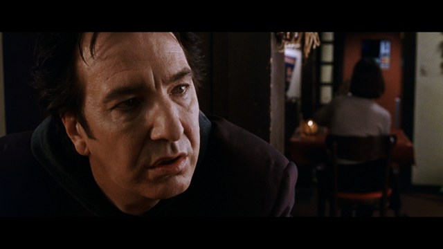 A close-up of a man with a concerned expression, with blurred figures in the background, set in a dimly lit interior.