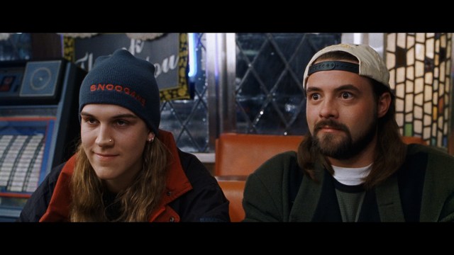 Two characters sitting in a diner, one wearing a blue beanie with 'SNOOGANS' and the other wearing a cap, both looking intently at something off-screen.