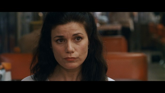 A woman with dark hair and a neutral expression sitting in a retro diner setting.