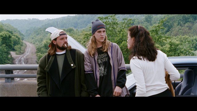A scene from a movie featuring three characters standing on a bridge with a lush green landscape in the background.