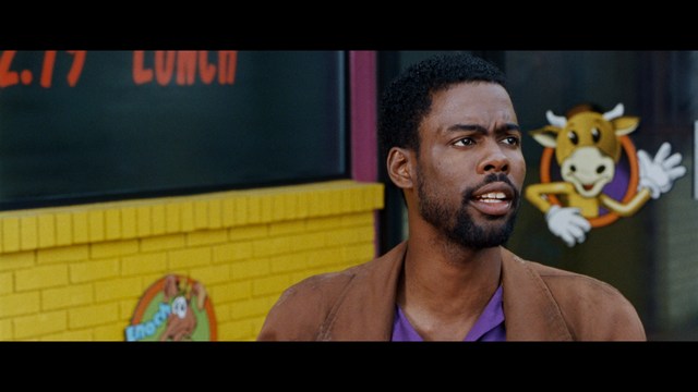 A scene featuring a man with short hair and a beard, wearing a brown jacket and purple shirt, standing outside a restaurant with bright yellow walls and a cartoon cow logo.