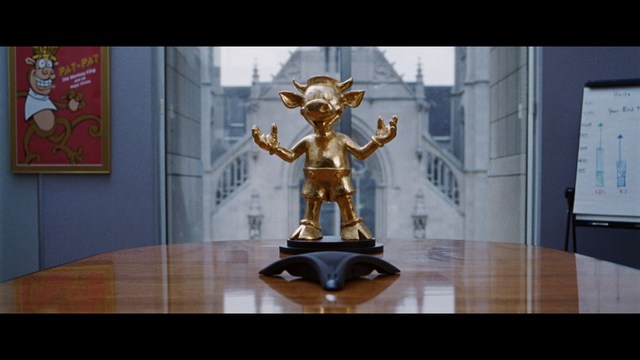 A golden trophy statue of a goblin-like character positioned on a wooden table in an office setting, with large windows and a building facade visible in the background.