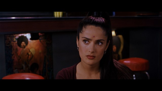 Close-up of a woman with long black hair sitting in a restaurant, looking concerned. A colorful painting is visible in the background, along with red bar stools.