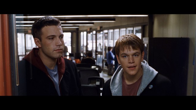 Two young men are looking at something off-camera, standing inside a building with bright lighting and seating areas visible in the background.
