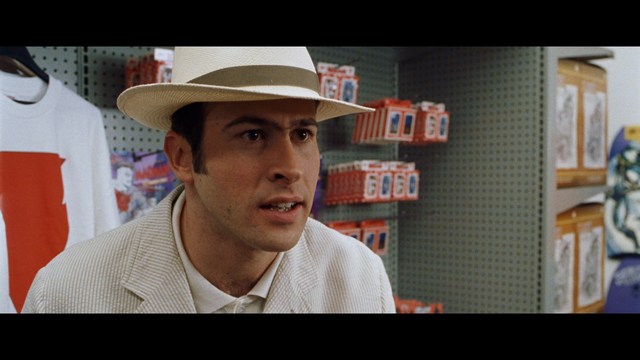A close-up shot of a man wearing a white suit and a white hat, looking intently at someone in a retail store filled with merchandise.