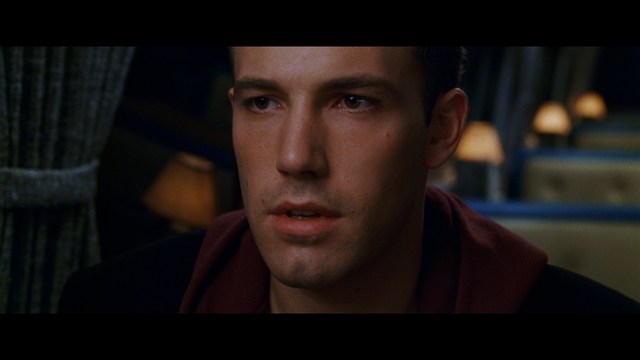 Close-up of a man with a serious expression, seated in a dimly lit area, with soft lighting from background lamps.