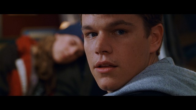 A close-up shot of a young man with short hair and a gray sweatshirt, looking intently at the camera, with a woman sleeping beside him in the background.