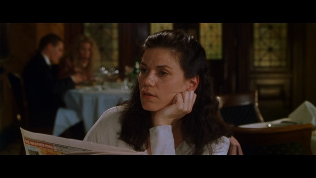 A woman seated at a restaurant table, thoughtfully resting her chin on her hand while reading a newspaper. Blurred figures of two people can be seen in the background.