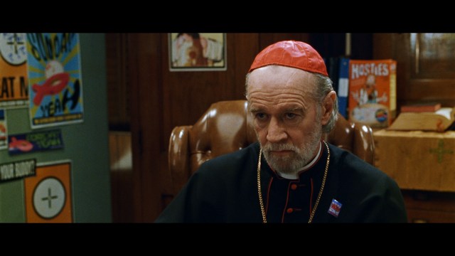 A serious-looking cardinal with a red hat and black robes sitting in a wooden chair in an office, surrounded by colorful posters and memorabilia.