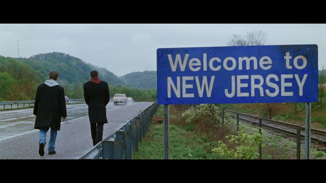 Two figures walking on a road towards a blue 'Welcome to New Jersey' sign.