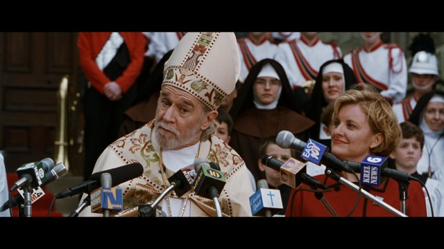 A scene featuring a man in a bishop's attire speaking at a podium surrounded by microphones, with a woman in a red dress beside him and a crowd of onlookers in the background.