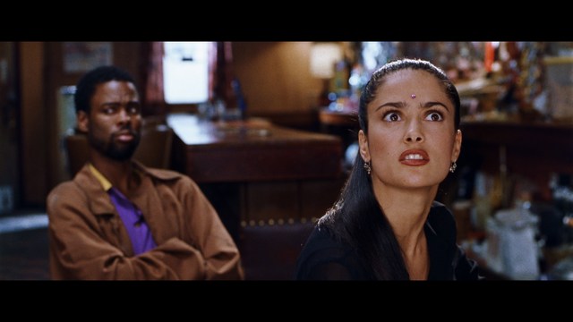 A tense scene featuring a woman with long hair and a serious expression, looking off to the side, while a man with short hair sits in the background, appearing relaxed and slightly turned away.