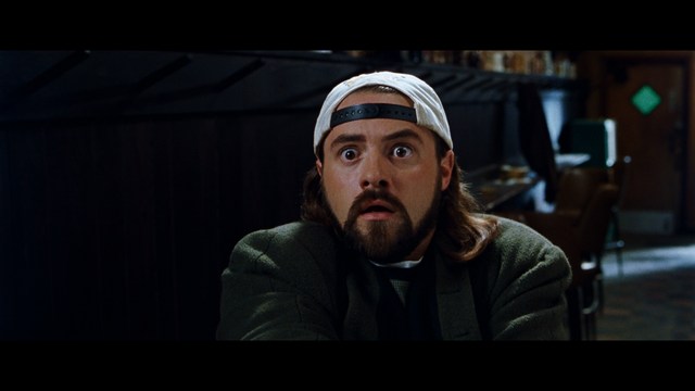 A shocked man with long hair and a baseball cap sitting in a bar, looking surprised.