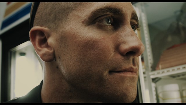 A close-up shot of a young man with short hair and a gray sweatshirt, looking intently at the camera, with a woman sleeping beside him in the background.