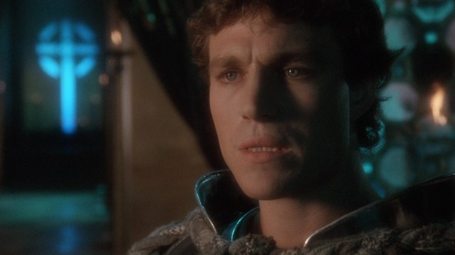 Close-up of a man with a serious expression, featuring details of his face and suit against a softly lit background.