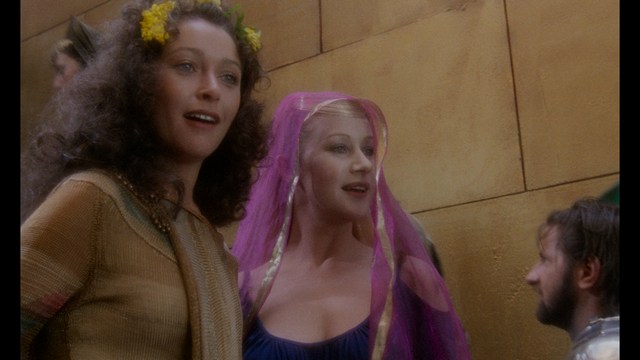 A tense scene featuring a woman with long hair and a serious expression, looking off to the side, while a man with short hair sits in the background, appearing relaxed and slightly turned away.