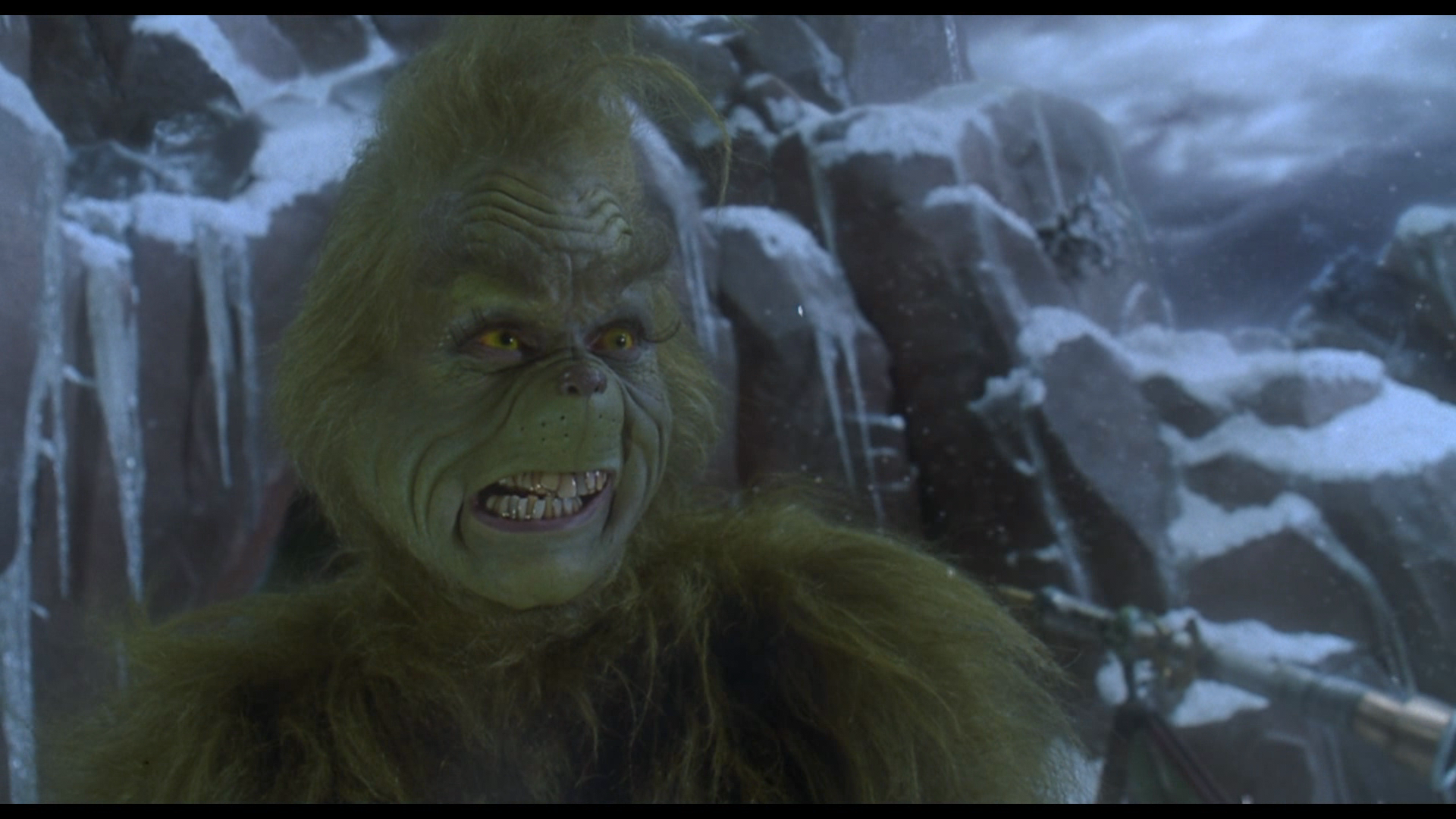 Close-up of a man with a serious expression, seated in a dimly lit setting, with another person in the background appearing to be seated at a table.