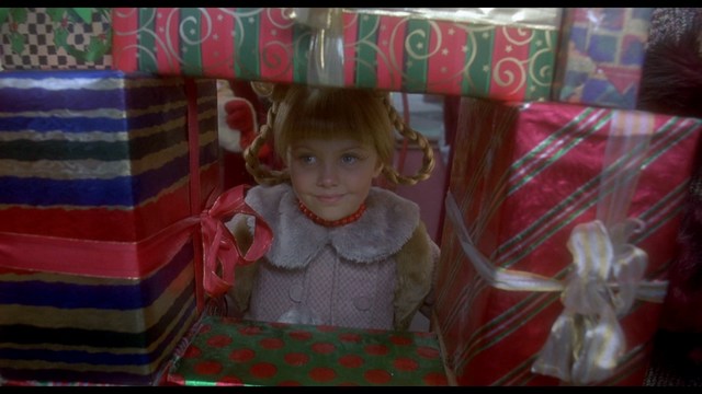 A man stands in front of an amusement arcade with a mural featuring a playful snowman and signs advertising games like skeeball, in a beach setting.