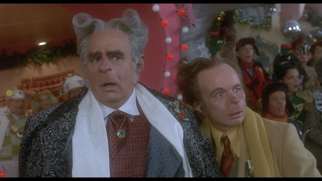 Two male characters sitting in a waiting area, one wearing a black jacket and the other in a maroon hoodie, with popcorn in hand.