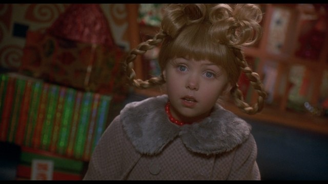 A woman with long dark hair, wearing a brown sweater, gestures with one hand while speaking in a warmly lit room with blinds and colorful artwork in the background.